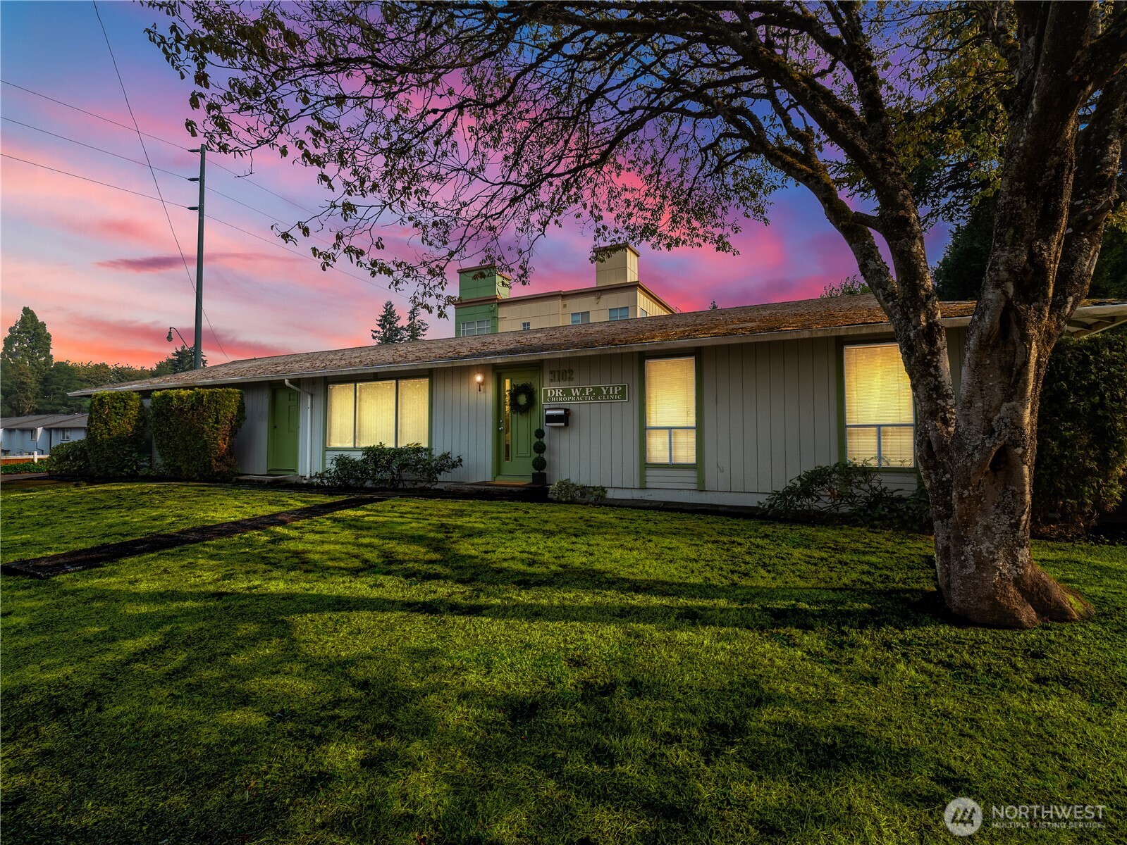 Single-story chiropractic clinic with green-trimmed windows, a broad lawn, and a pink sunset.