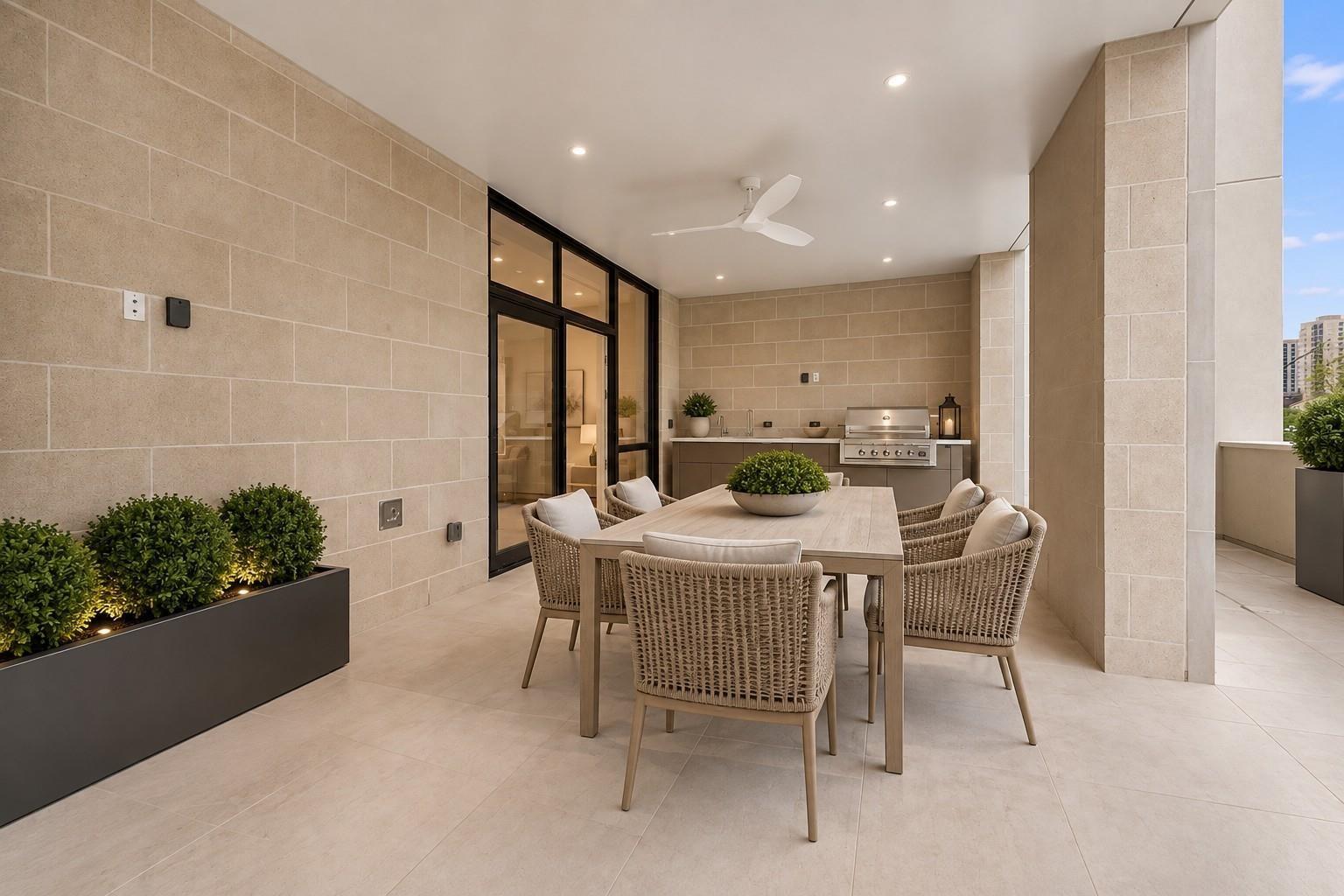 Covered stone patio with a dining table, woven chairs, potted greenery, and built-in grill.