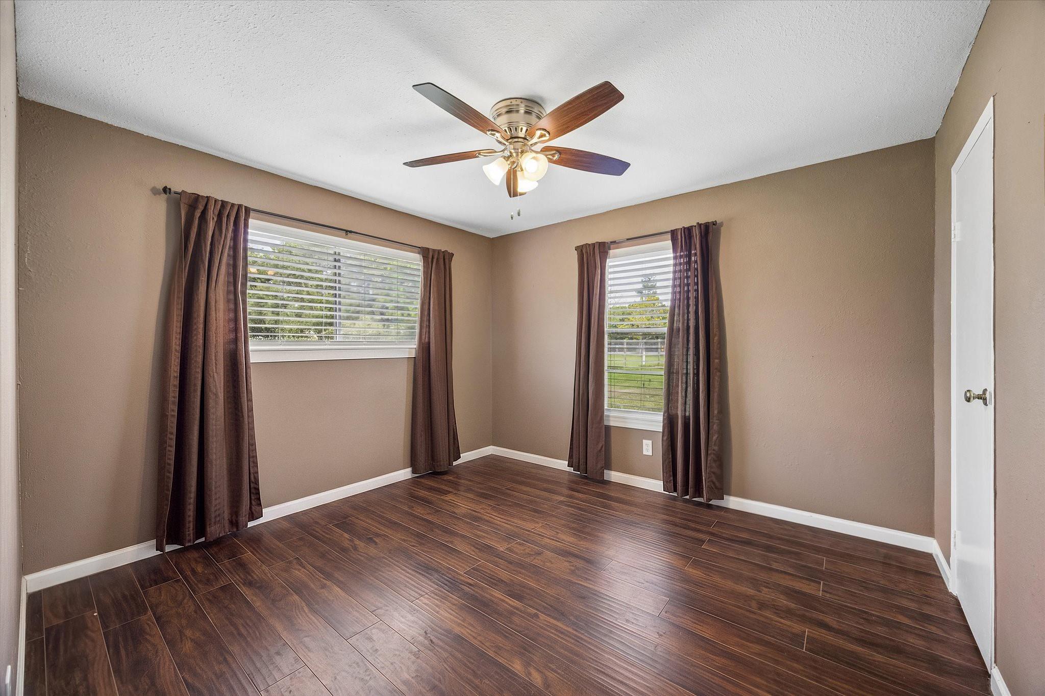 Empty bedroom with dark wood-look floors, two curtained windows, and a ceiling fan.