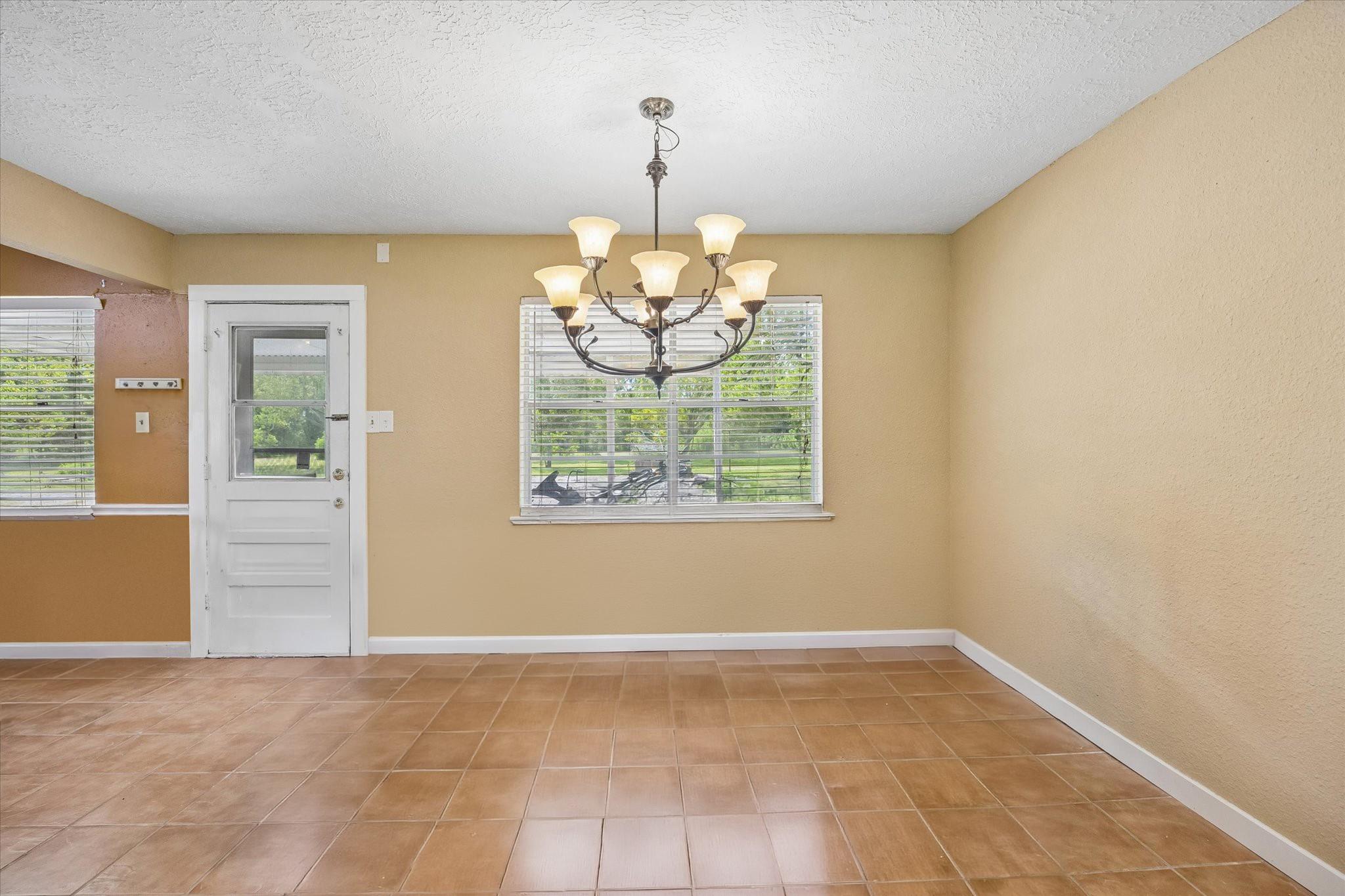 Empty dining room with tan walls, brown tile flooring, and a chandelier above the windows.