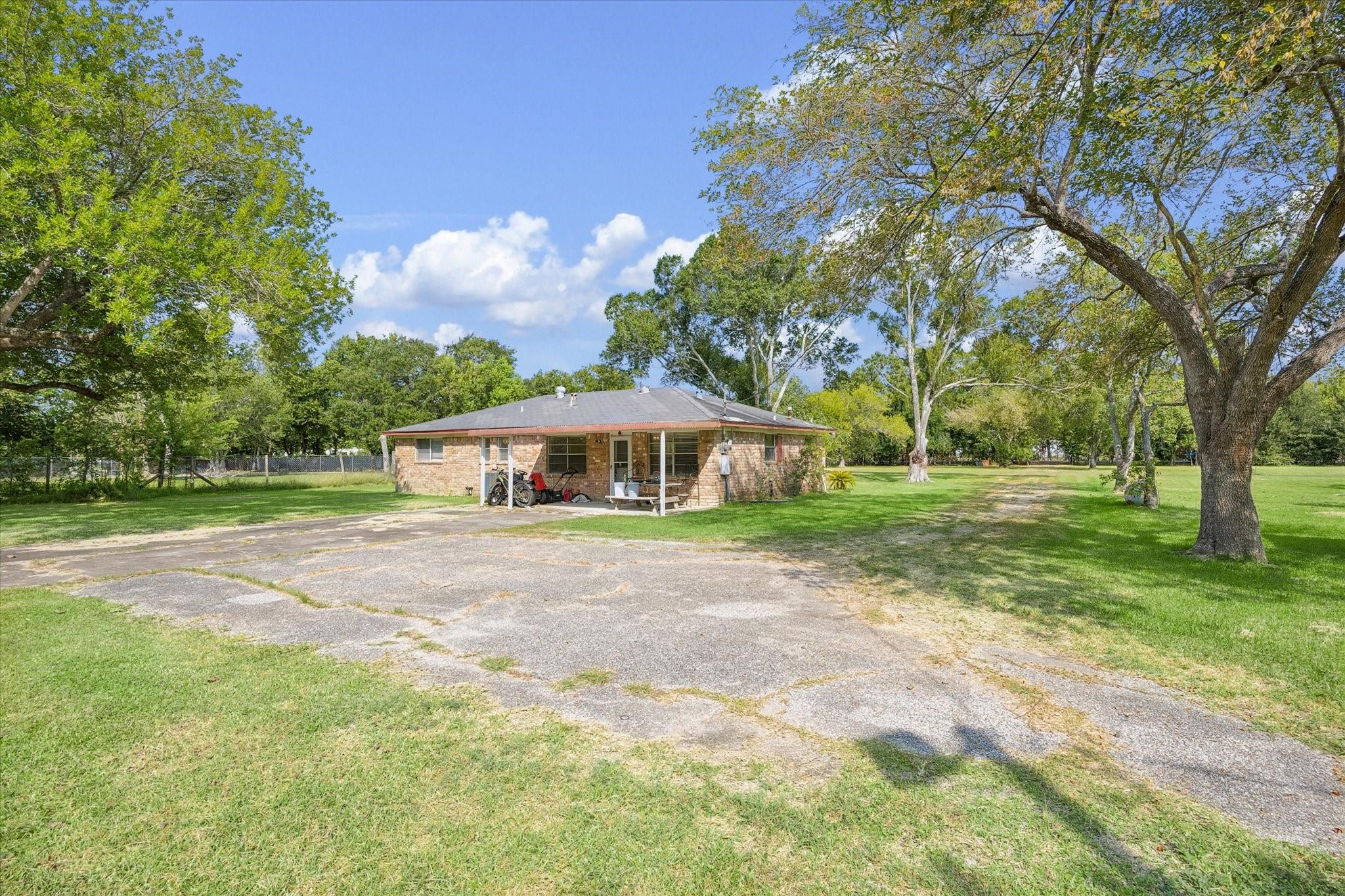 Single-story brick house with a covered patio, broad paved driveway, and mature trees across a grassy lot.