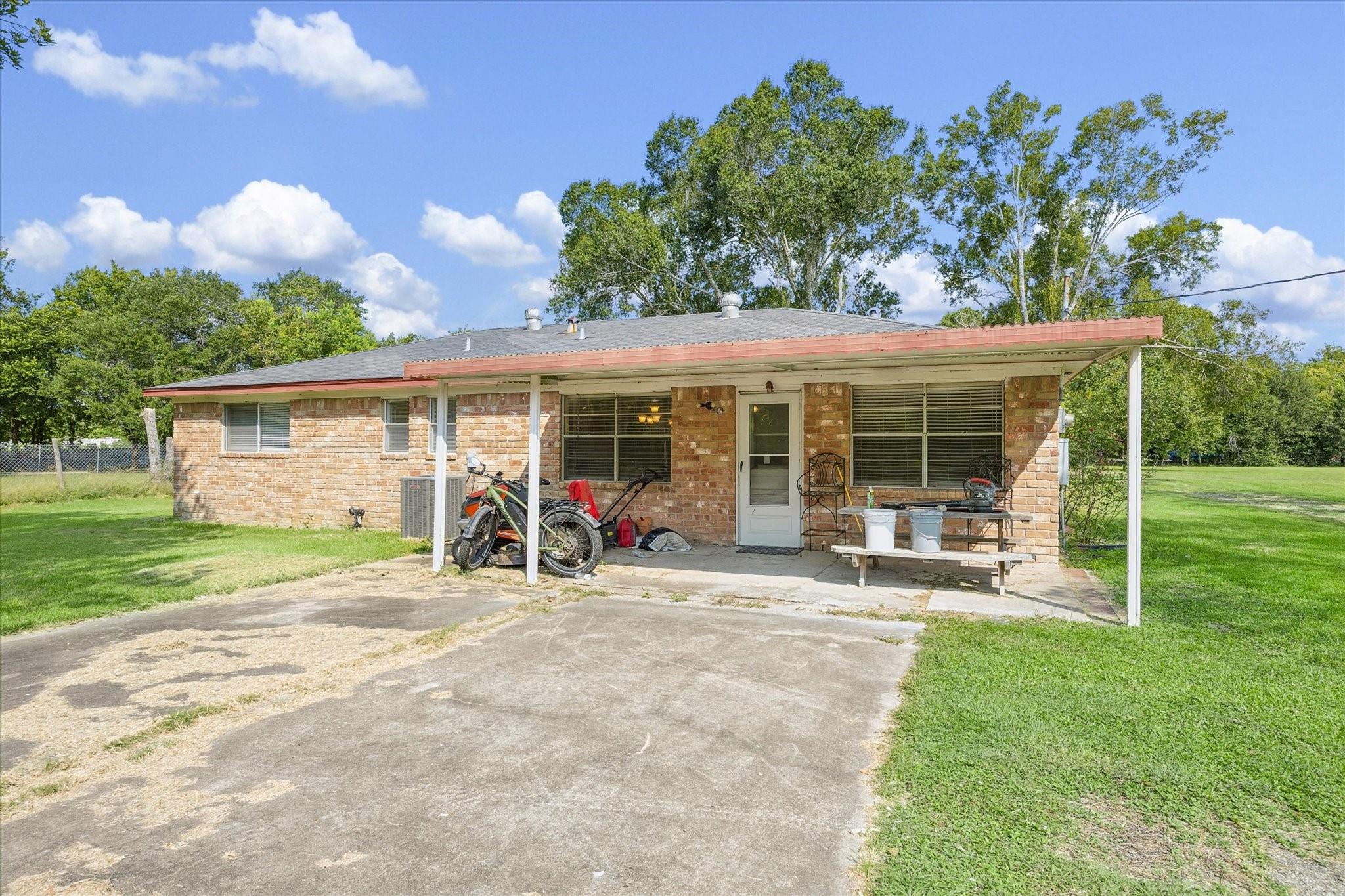 Single-story brick home with a broad covered patio, concrete driveway, and mature trees.