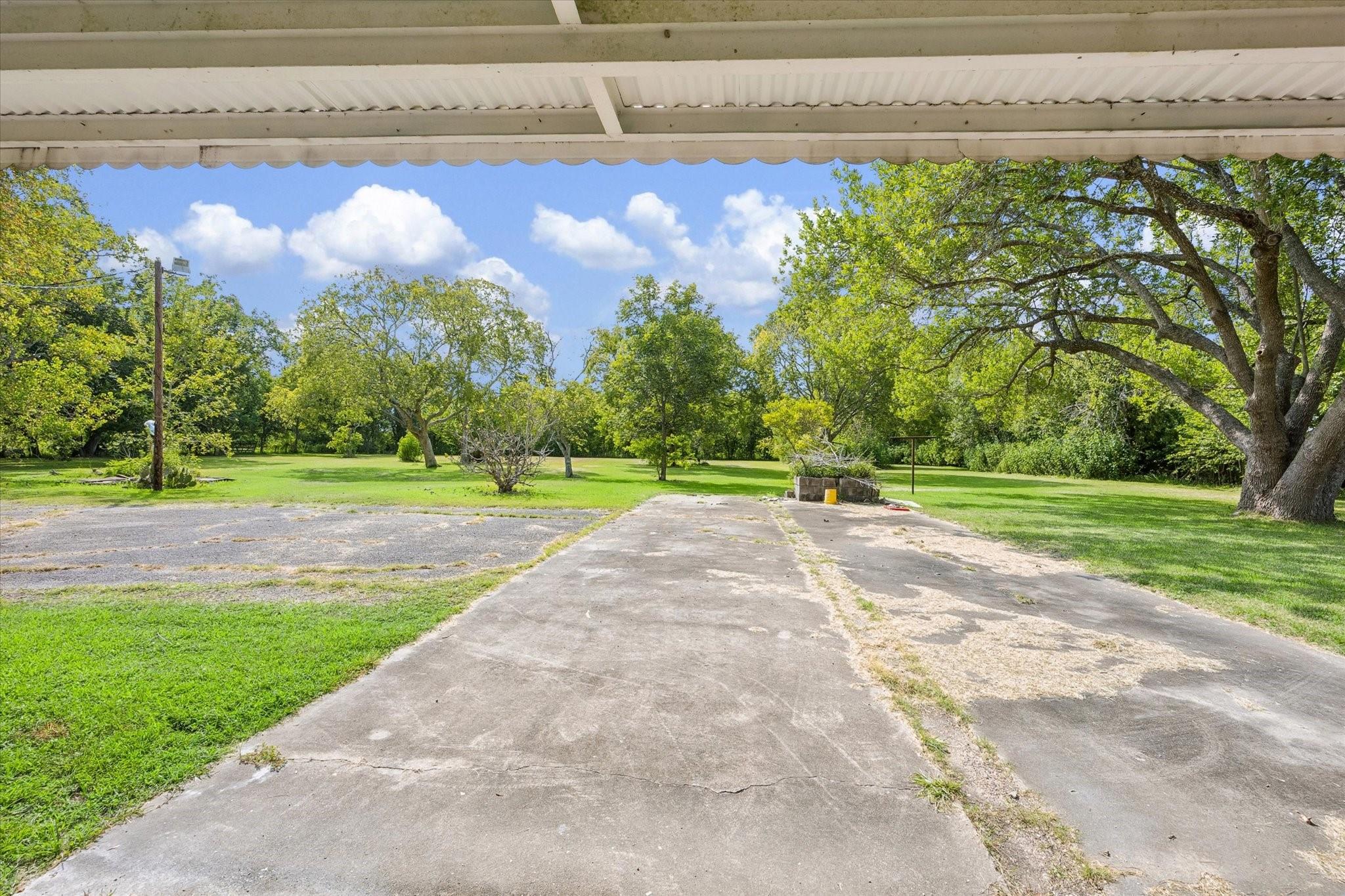 Concrete driveway beneath a carport opens onto a tree-shaded lawn.