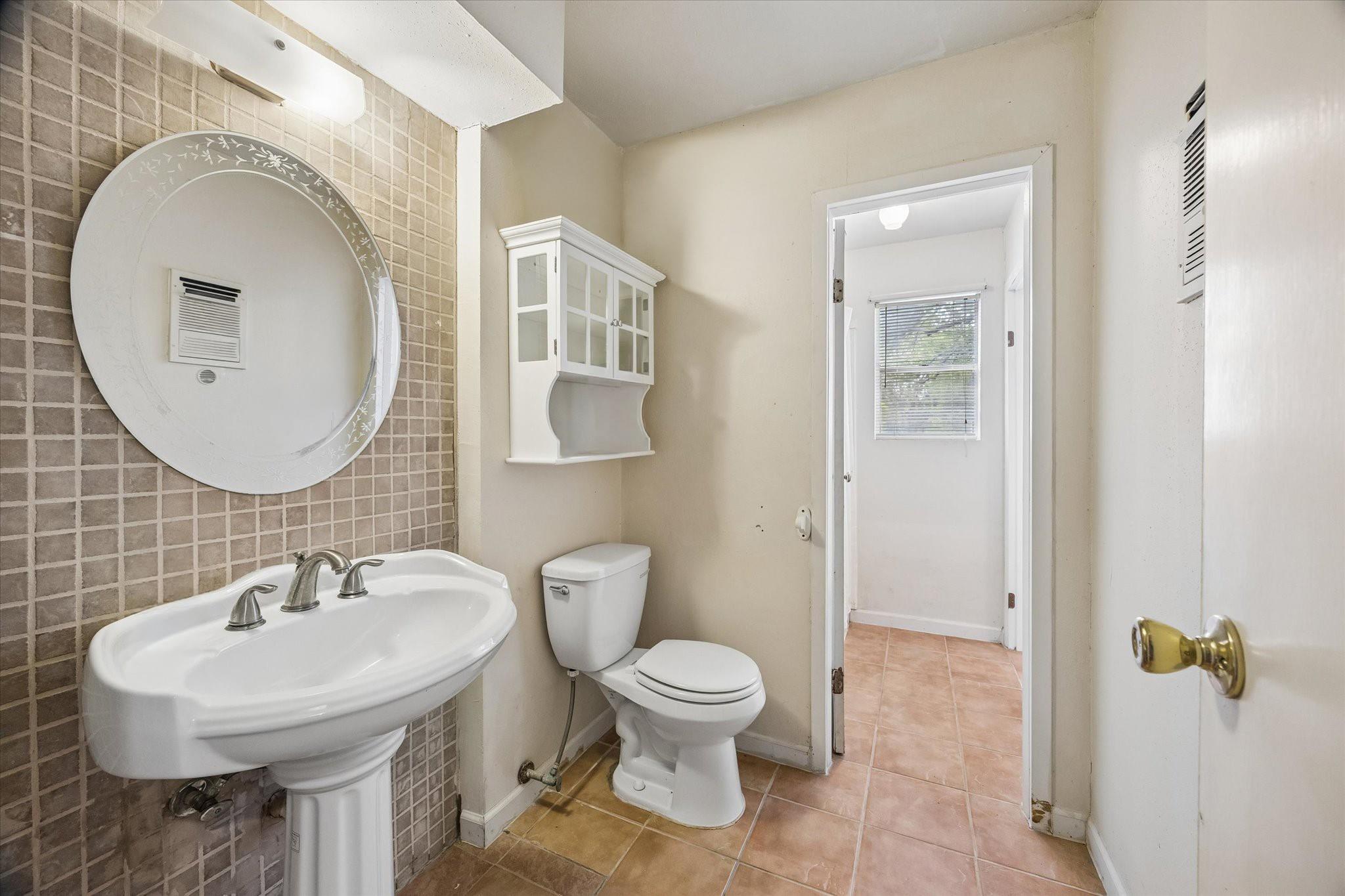 Bathroom with pedestal sink, round mirror, tiled wall, toilet, and open doorway.