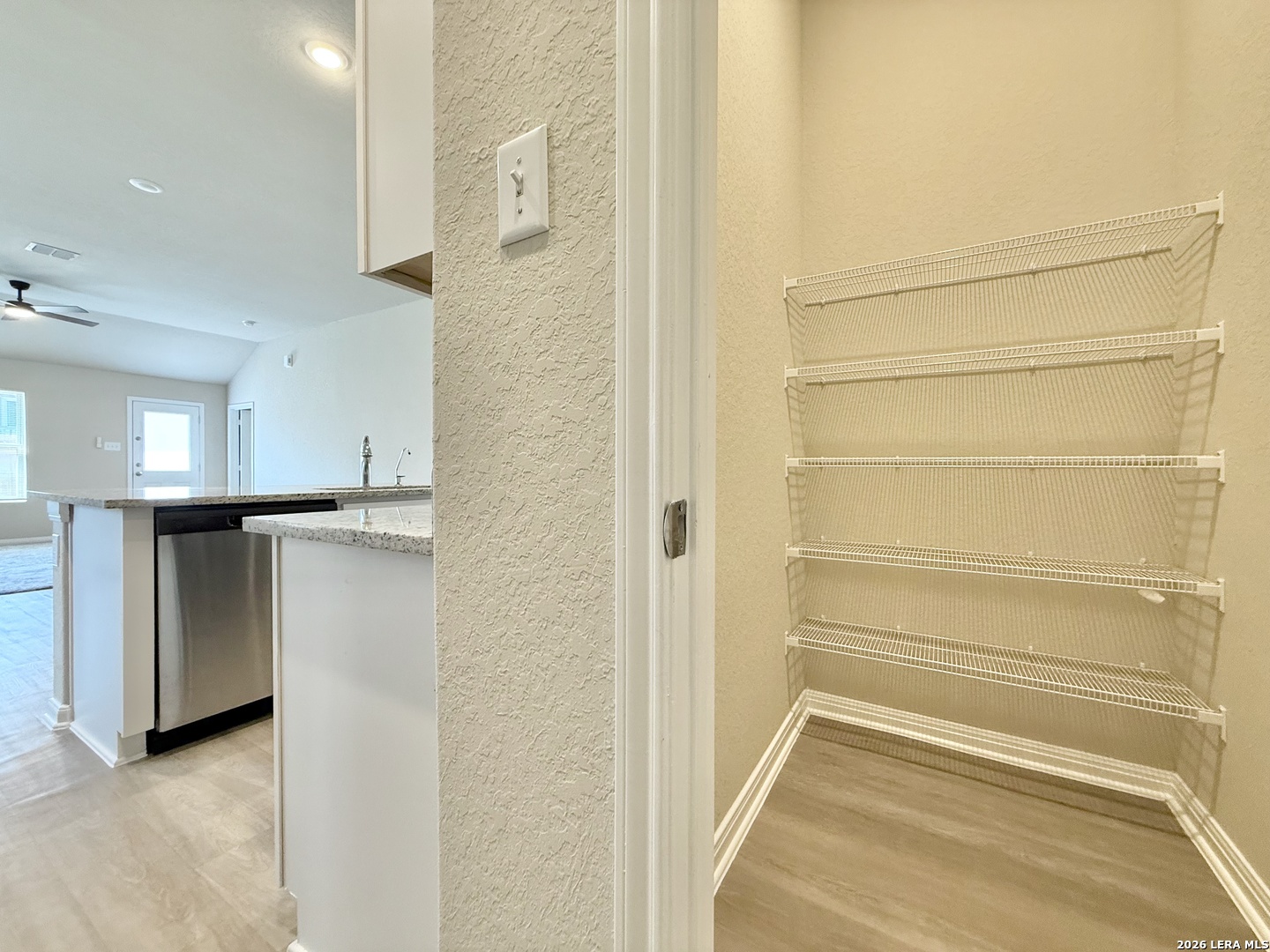 Walk-in pantry with white wire shelves beside a kitchen with granite counters and a stainless-steel dishwasher.