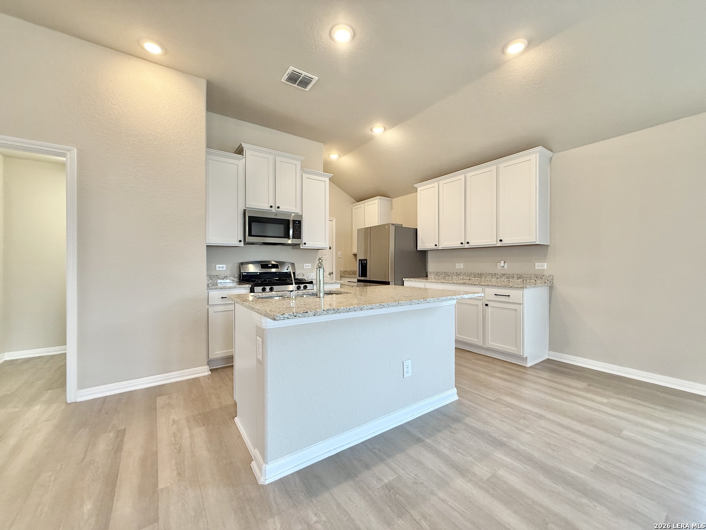 Kitchen with white cabinets, granite island and sink, stainless steel appliances, and light wood-look flooring.