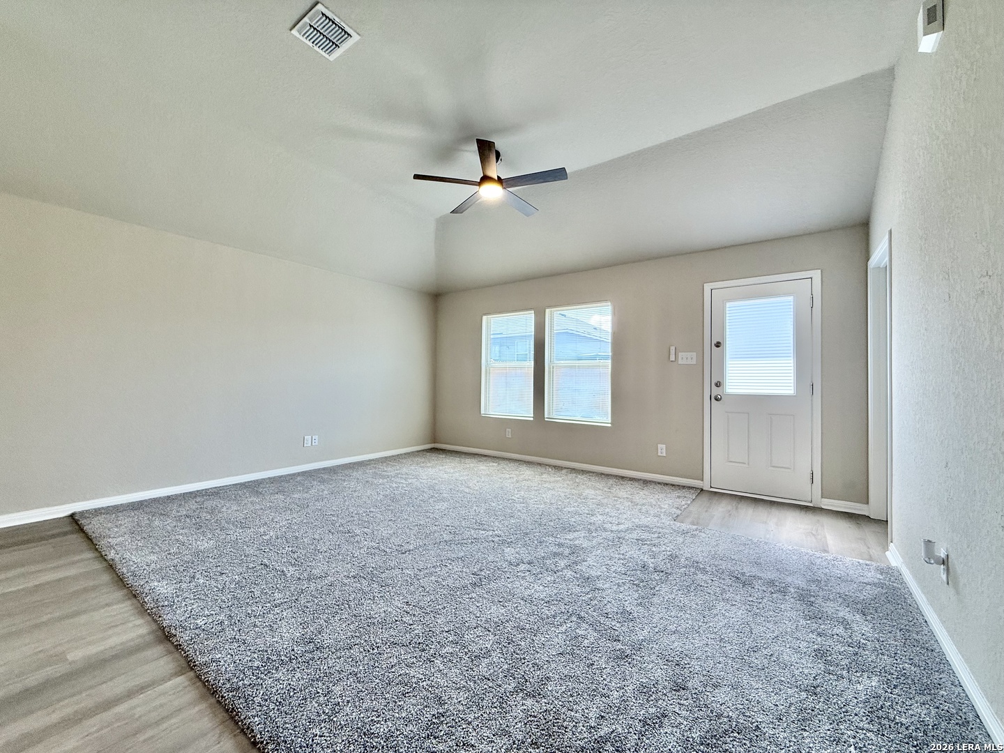 Vacant living room with gray carpet, vaulted ceiling, ceiling fan, two windows, and a glazed exterior door.