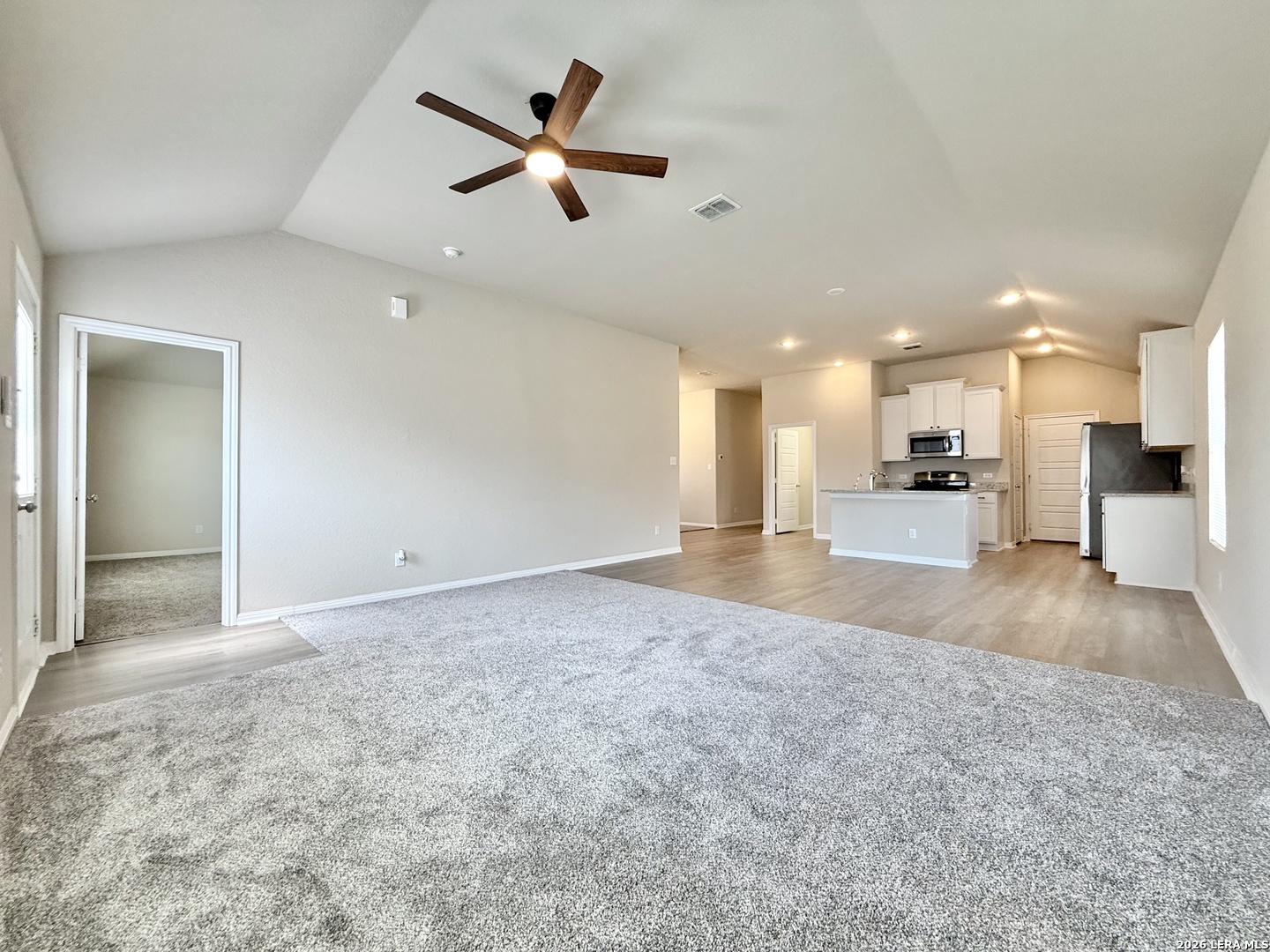 Vacant open-plan living room and kitchen with gray carpet, a kitchen island, and a wood-blade ceiling fan.
