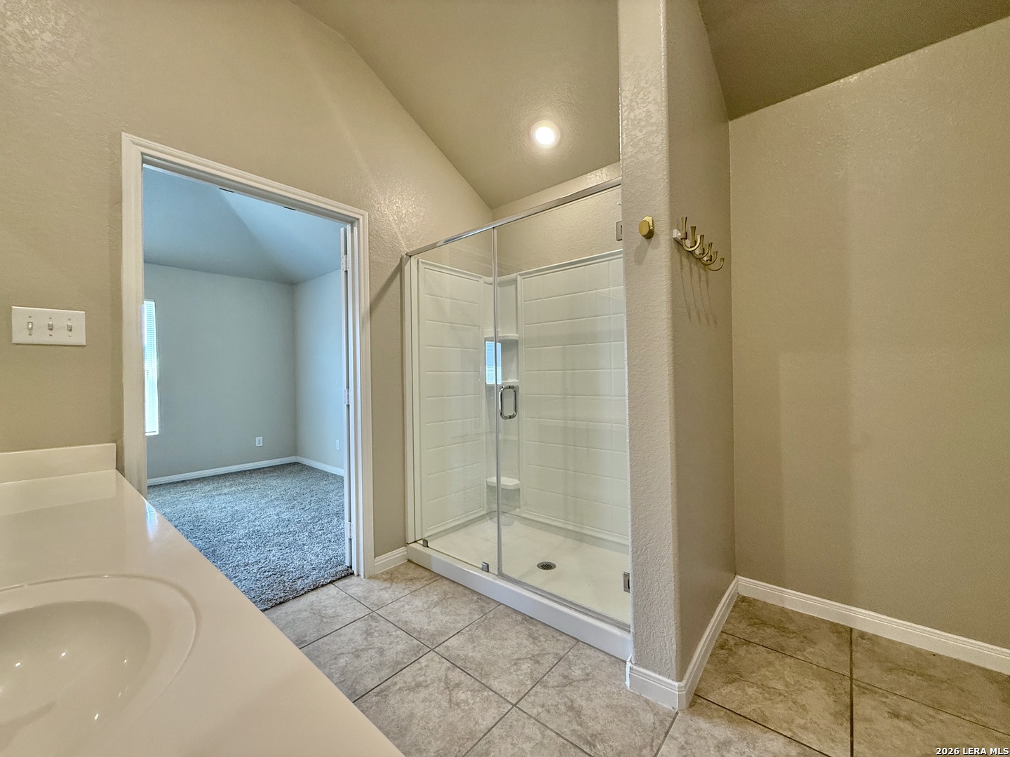 Bathroom with a glass shower and tiled floor, opening onto a carpeted adjoining room.