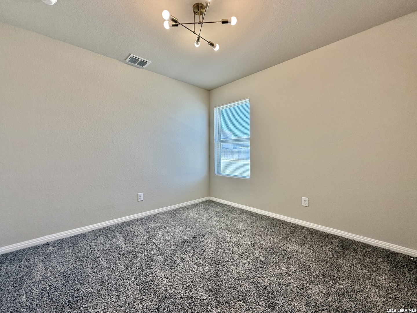 Vacant bedroom with gray patterned carpet, a window with blinds, and a modern ceiling light.