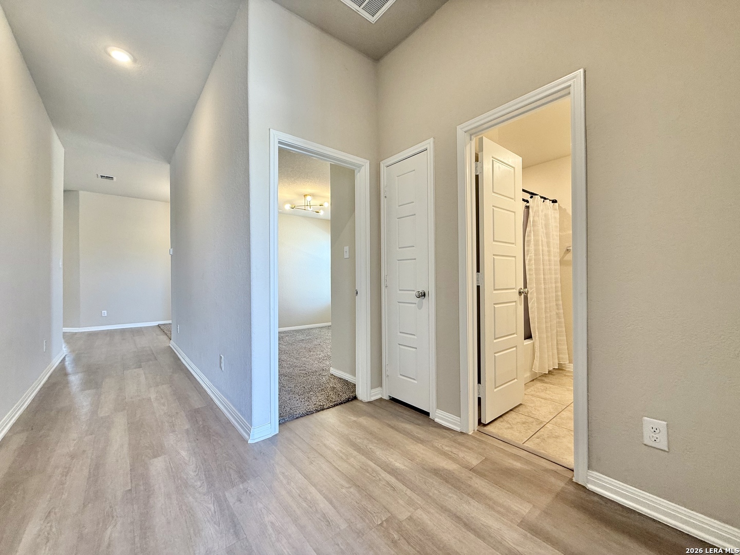 Empty hallway with light wood-look flooring, a carpeted room, and an open bathroom doorway.