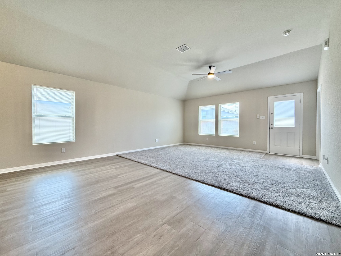 Empty living room with vaulted ceiling, ceiling fan, wood-look flooring, and a large carpeted area.
