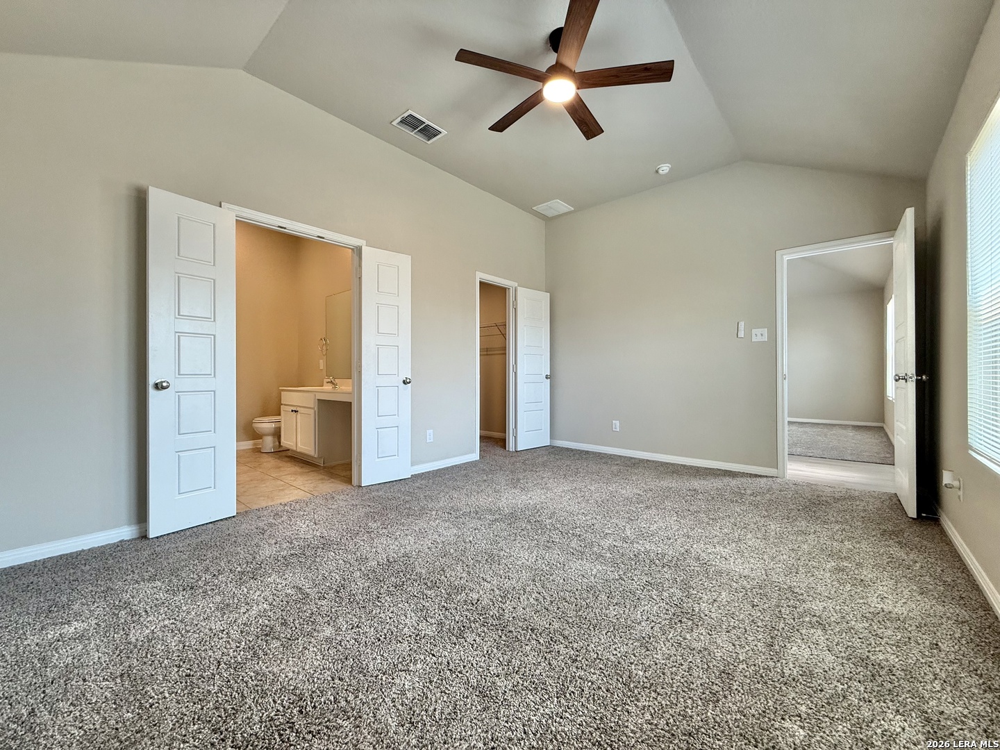 Vacant bedroom with gray carpet, vaulted ceiling, ceiling fan, and adjoining bathroom and closet.
