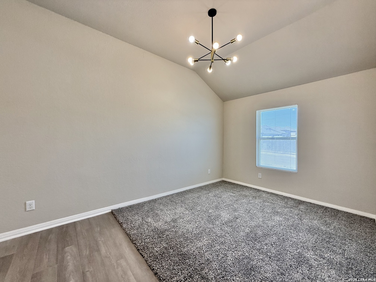 Empty bedroom with vaulted ceiling, gray carpet, window blinds, and a modern chandelier.