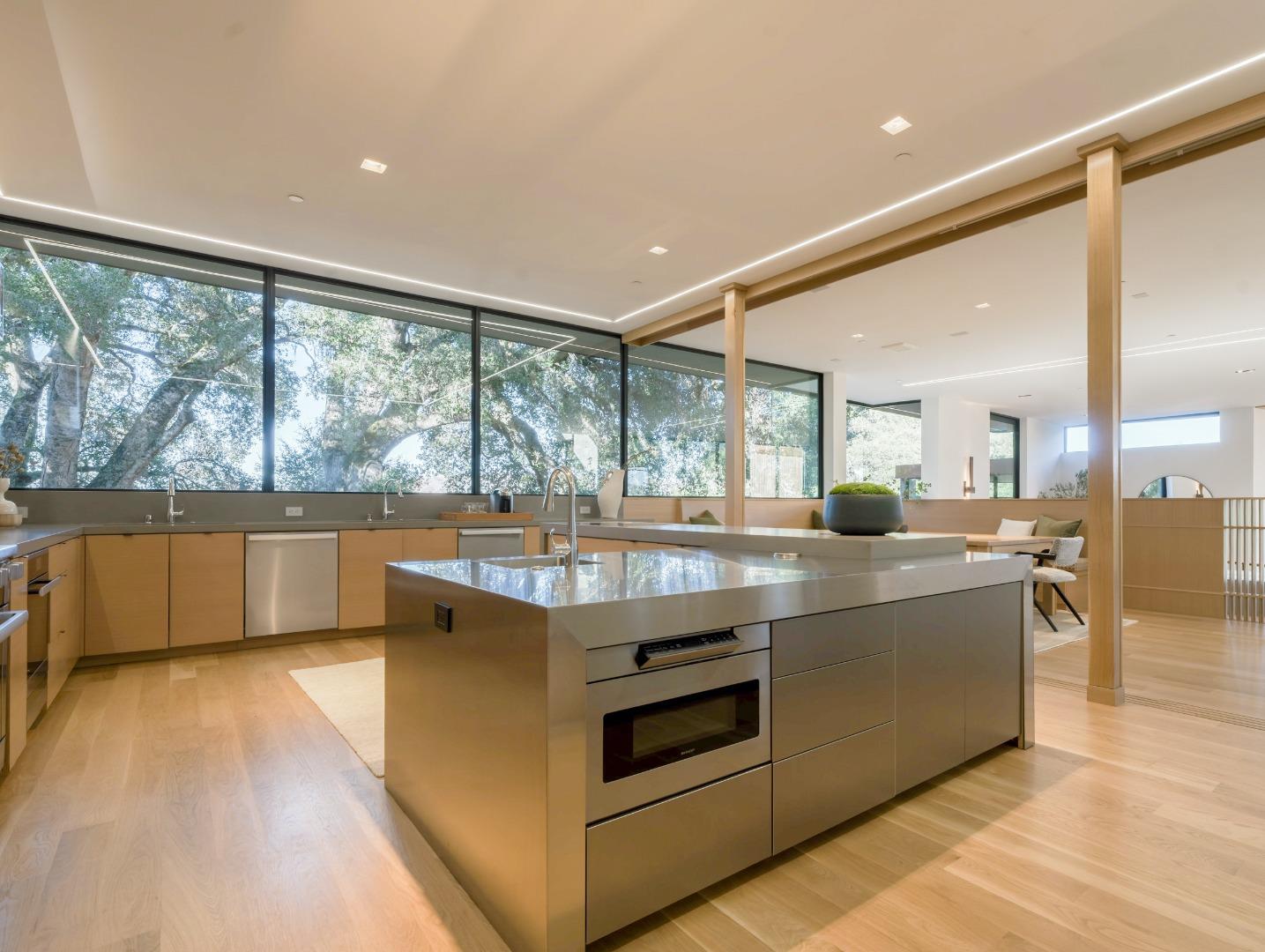 Modern kitchen with a large island, light wood cabinetry, and windows overlooking trees.