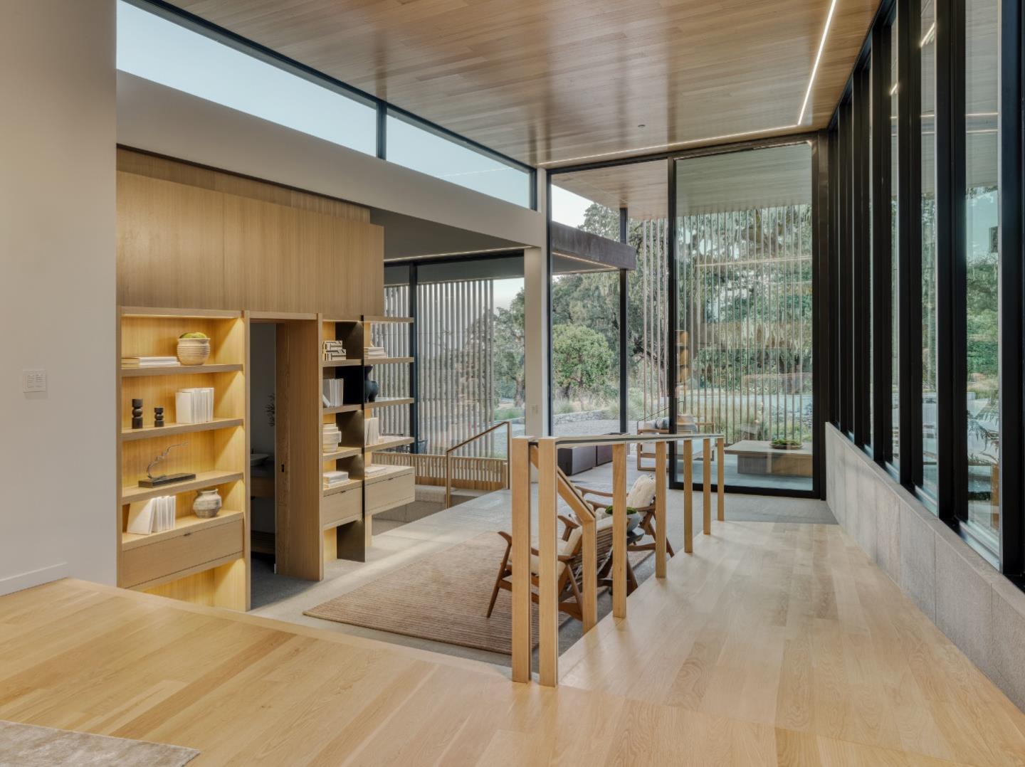 Modern living area with illuminated built-in shelves and floor-to-ceiling windows overlooking trees.