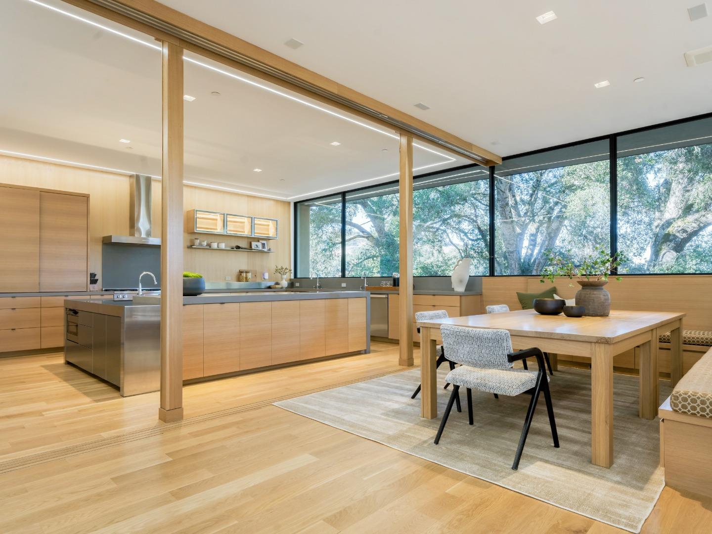 Modern oak kitchen and dining area with a large island and leafy trees visible through expansive windows.