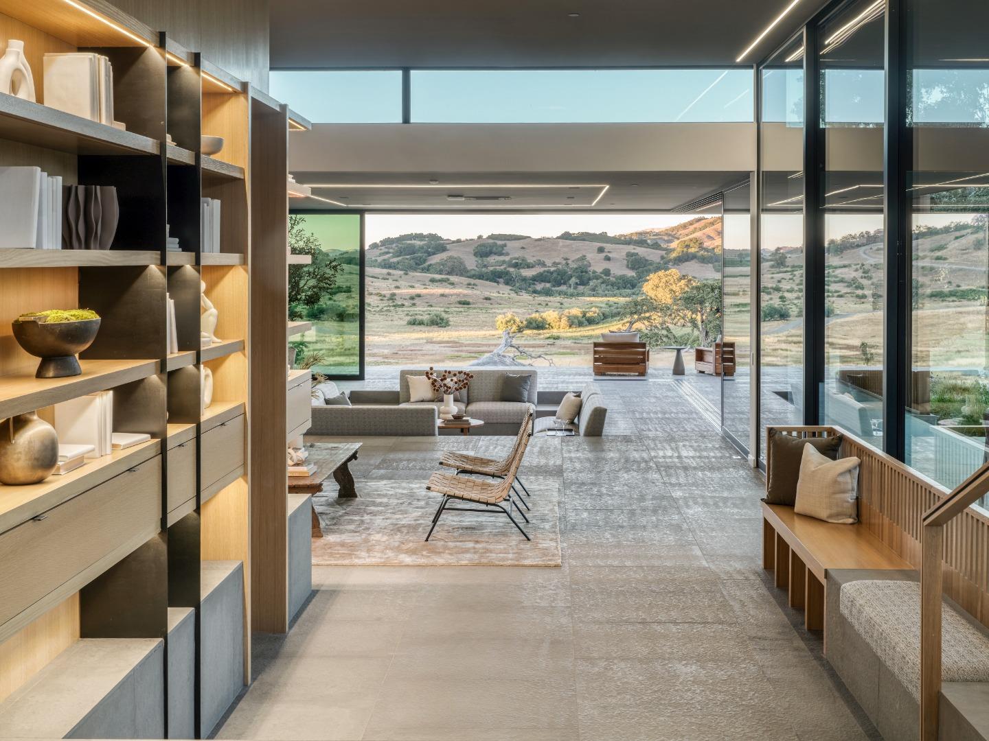 Modern living room with floor-to-ceiling glass overlooking rolling hills and warm-lit built-in shelving.