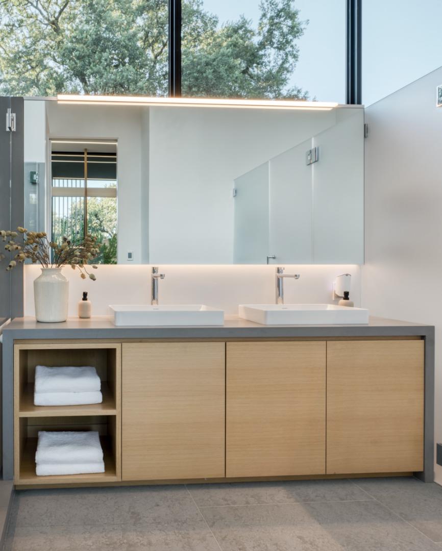 Modern bathroom vanity with two vessel sinks, light-wood cabinetry, and a large mirror beneath a tall window.