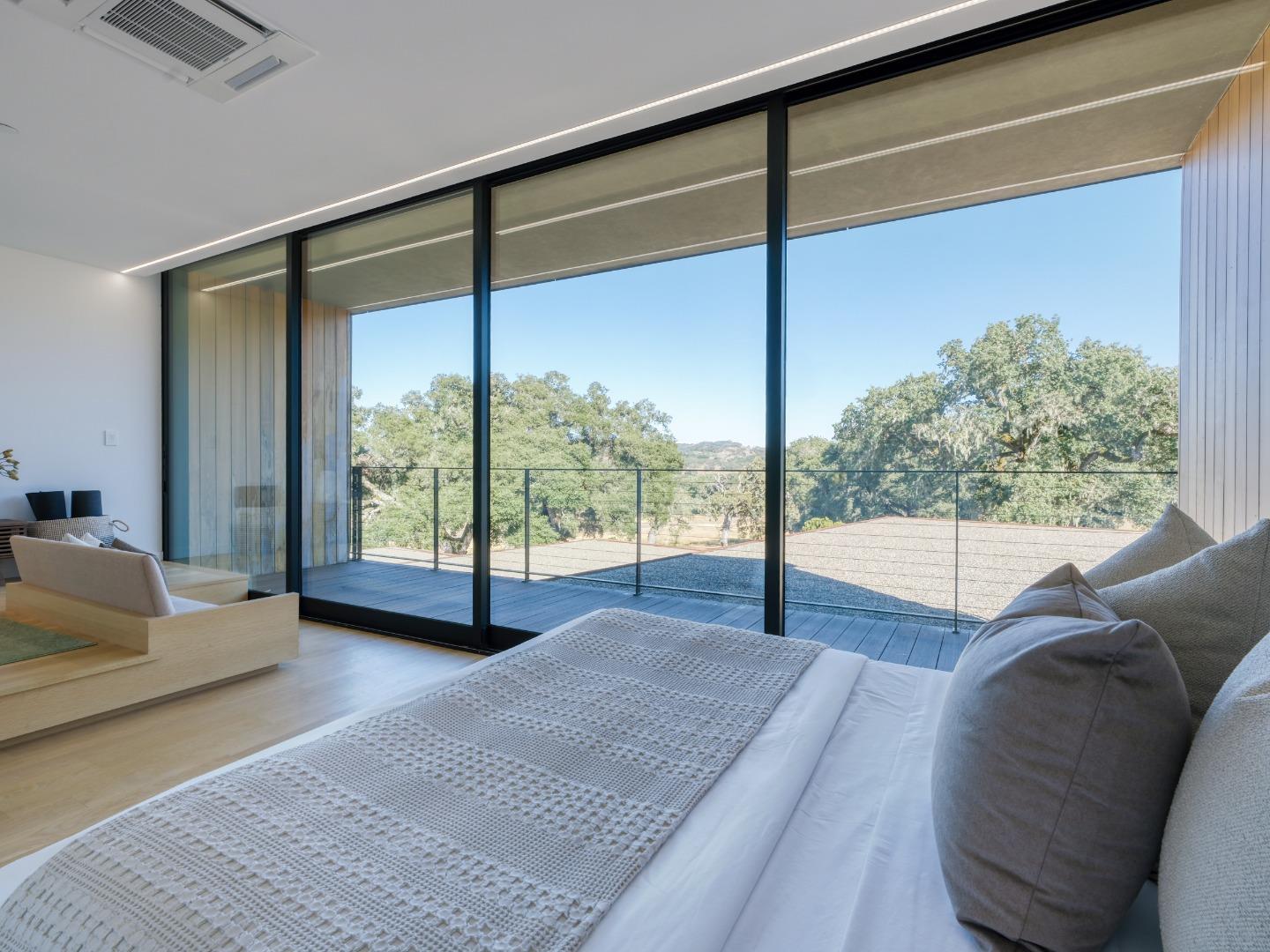 Modern bedroom with floor-to-ceiling glass doors opening to a balcony overlooking trees and hills.