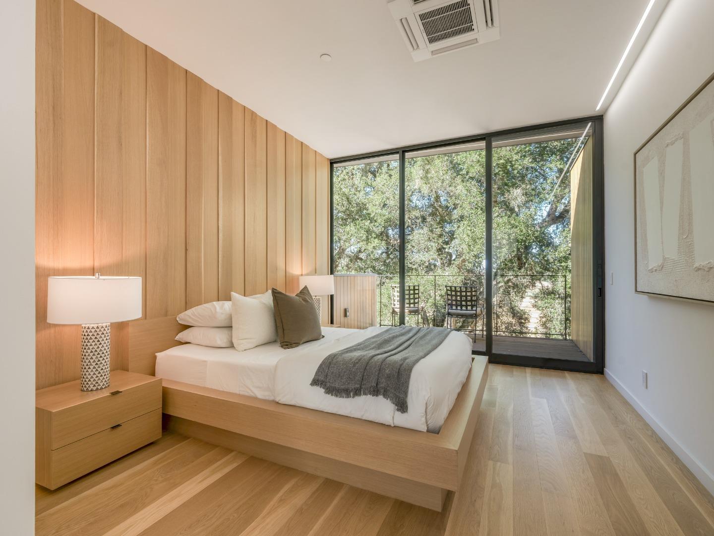 Modern bedroom with a low wood platform bed and floor-to-ceiling doors opening to a tree-lined balcony.