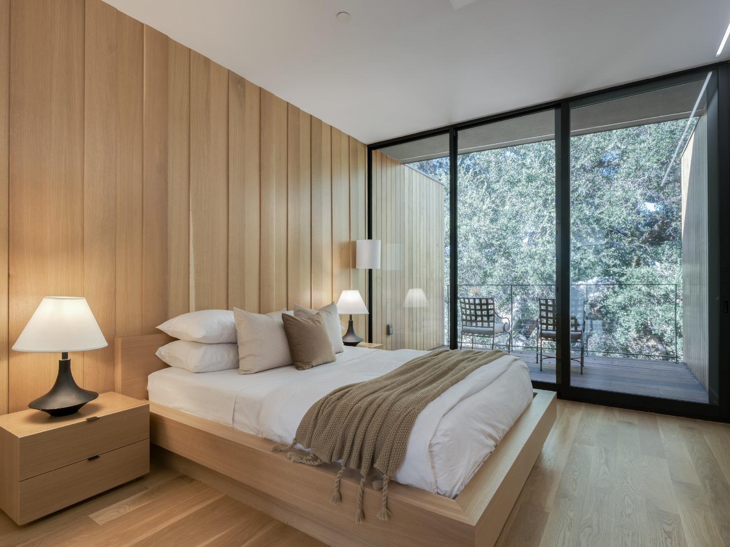 Modern bedroom with a wood platform bed, paneled wall, and glass doors overlooking trees.