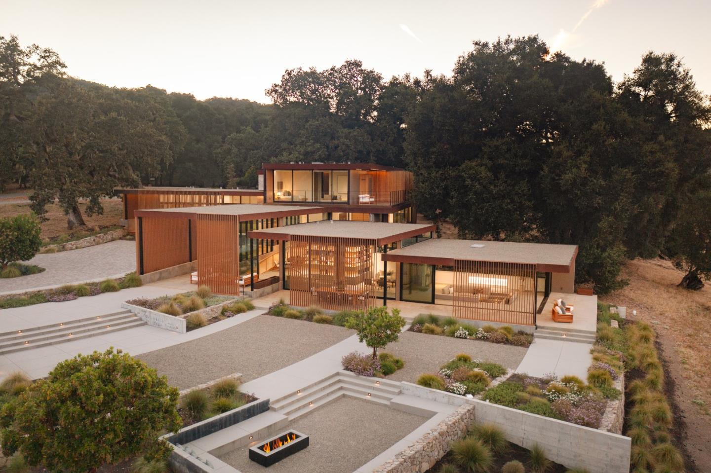 Modern wood-and-glass home with terraced landscaping and a sunken fire pit, surrounded by trees at sunset.