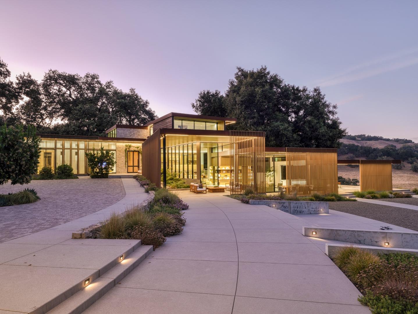 Modern glass-and-stone home with illuminated wood screens and a broad landscaped entry at dusk.