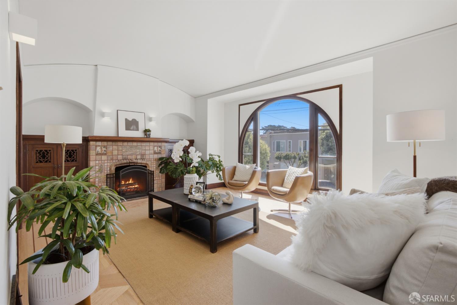 Staged living room with a tiled fireplace, arched window, and cream seating.