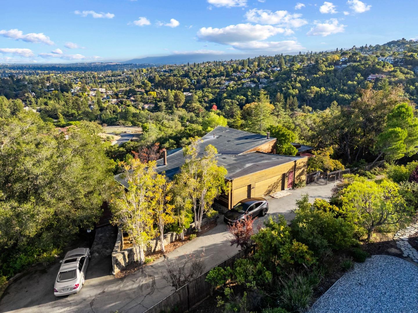 Aerial view of a wood-sided hillside home amid dense trees, with a wide driveway and valley beyond.