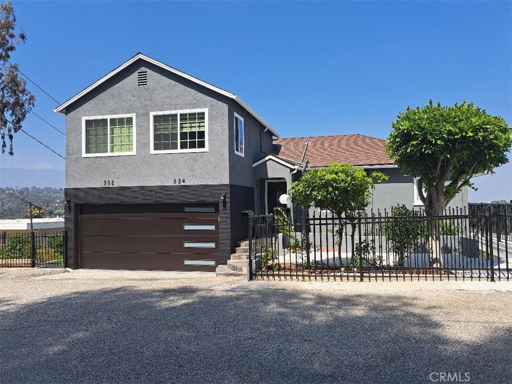 Gray two-story house with a dark garage door, black metal fence, and leafy front-yard trees.