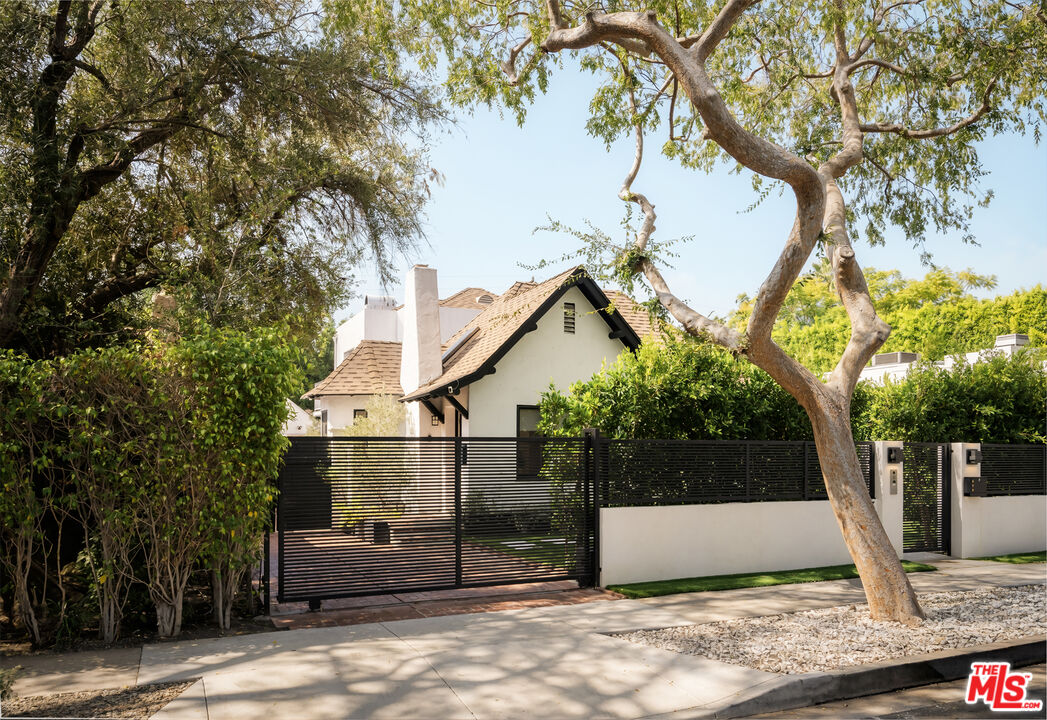White stucco house behind a black horizontal-slat gate, framed by mature trees.