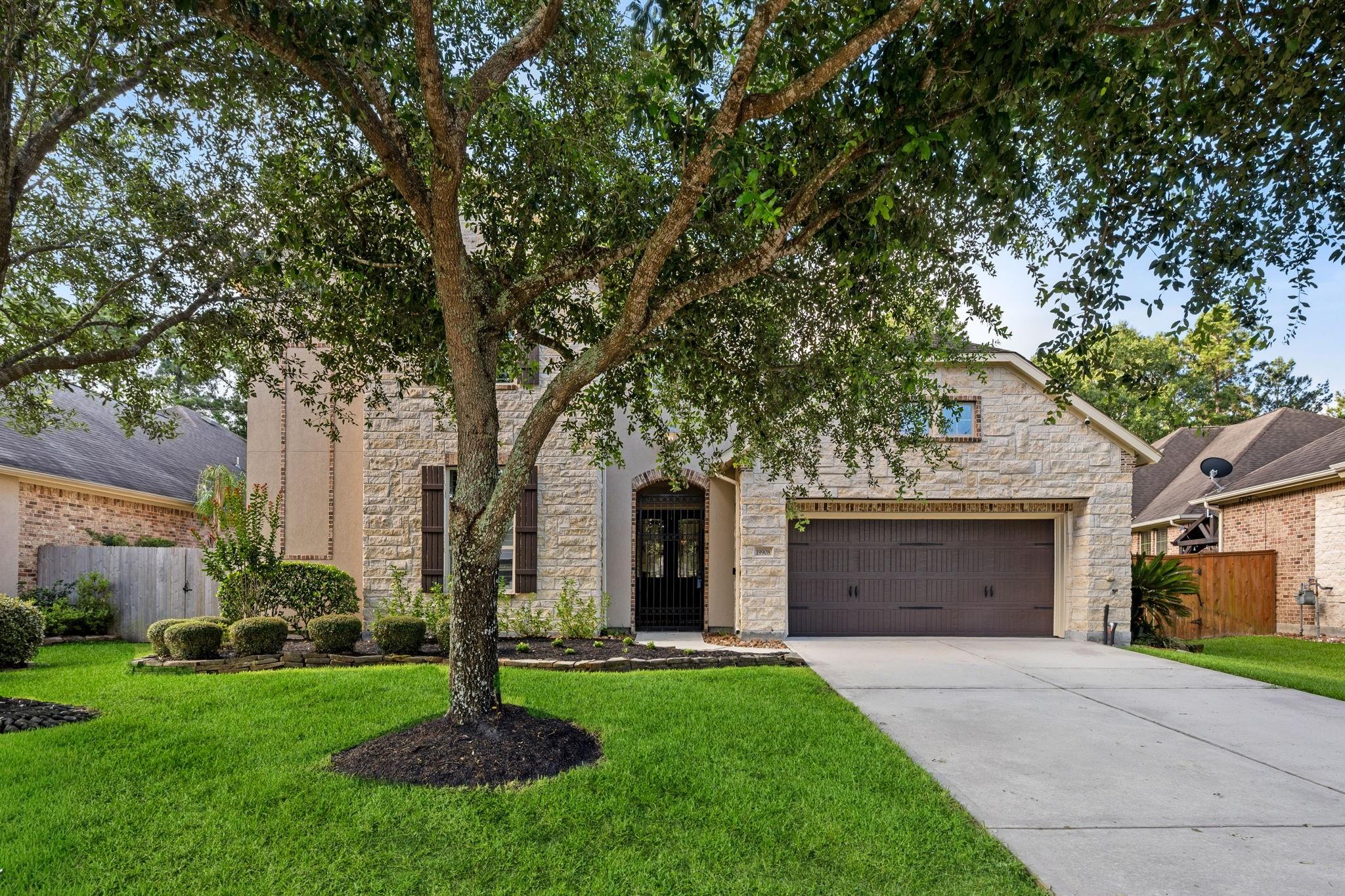 Two-story stone-front home with a mature shade tree, landscaped lawn, and double garage.