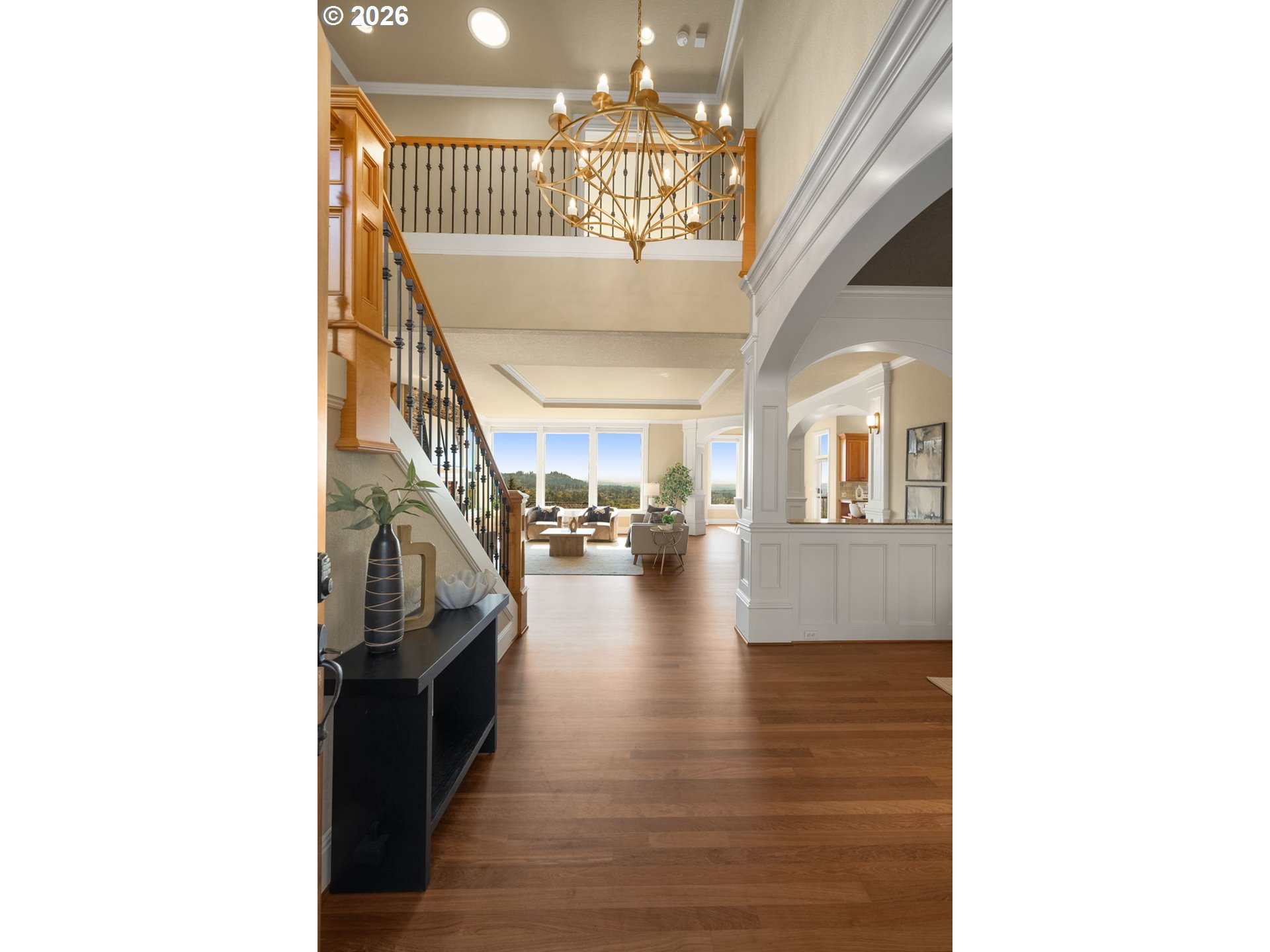 Furnished entry hall with a dramatic gold chandelier, open staircase, and living room windows overlooking wooded hills.