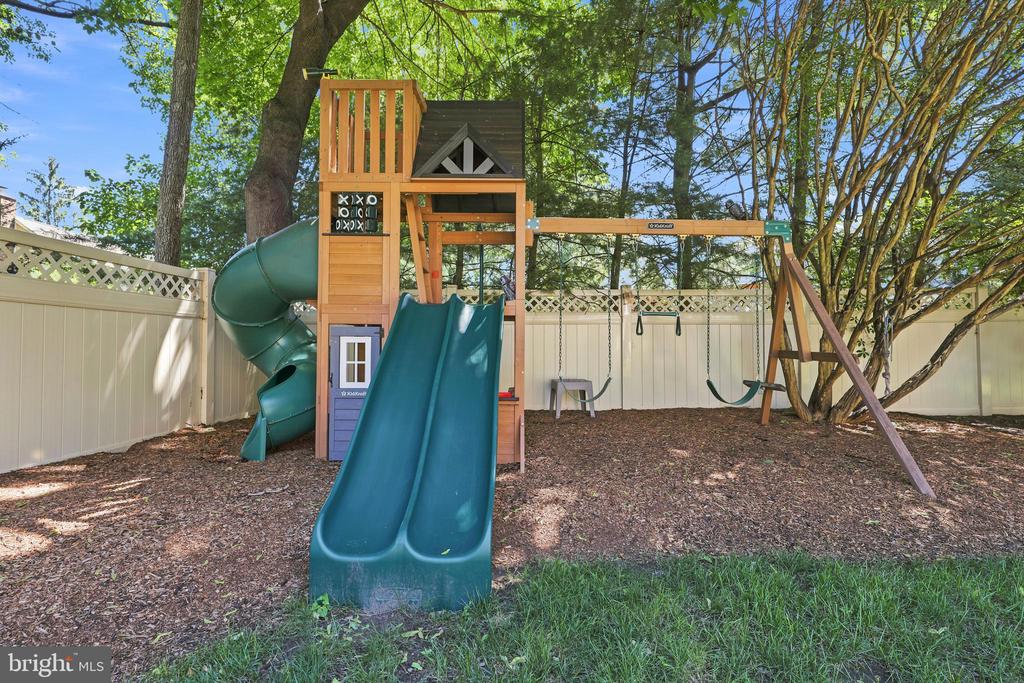Fenced backyard with a wooden playset, teal slides, and swings beneath mature trees.