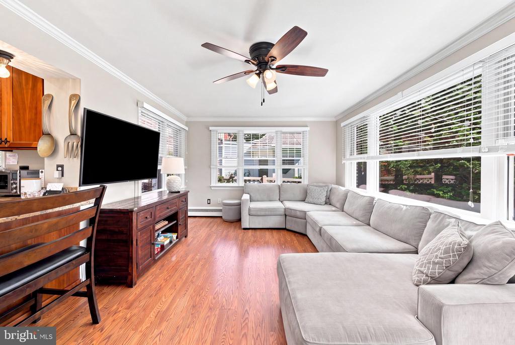 Living room with a large gray sectional, wood floors, ceiling fan, and wide windows overlooking greenery.