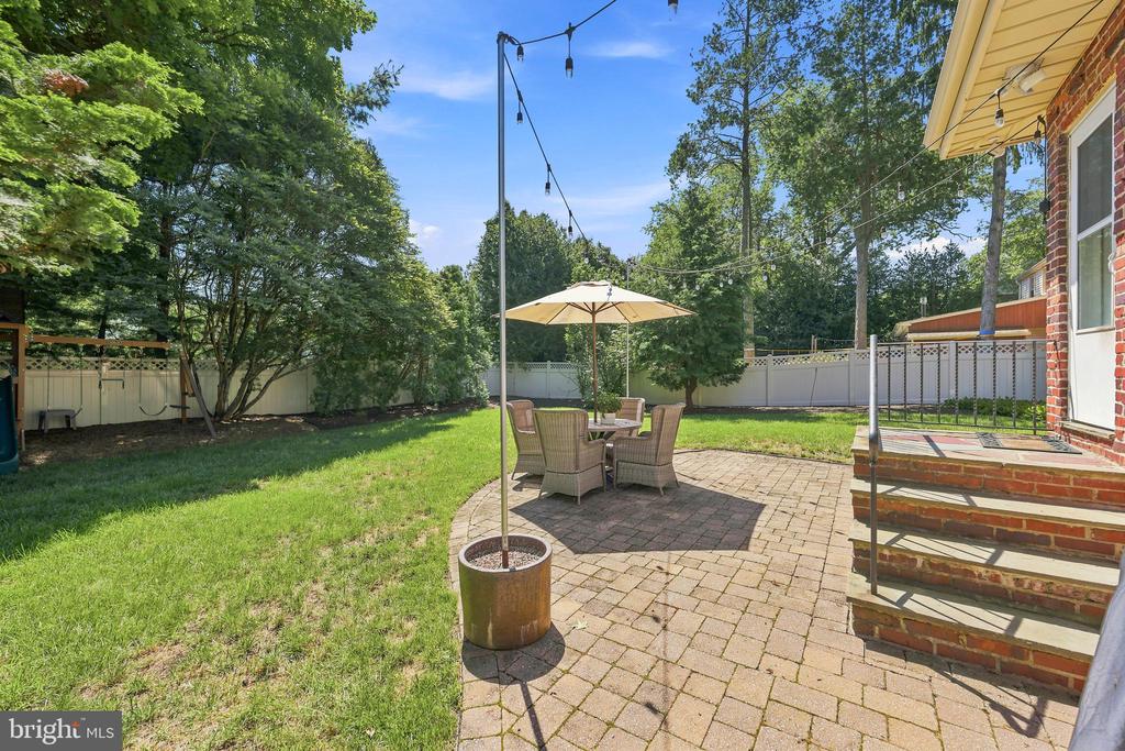 Backyard patio with brick pavers, umbrella-shaded chairs, lawn, mature trees, and string lights.