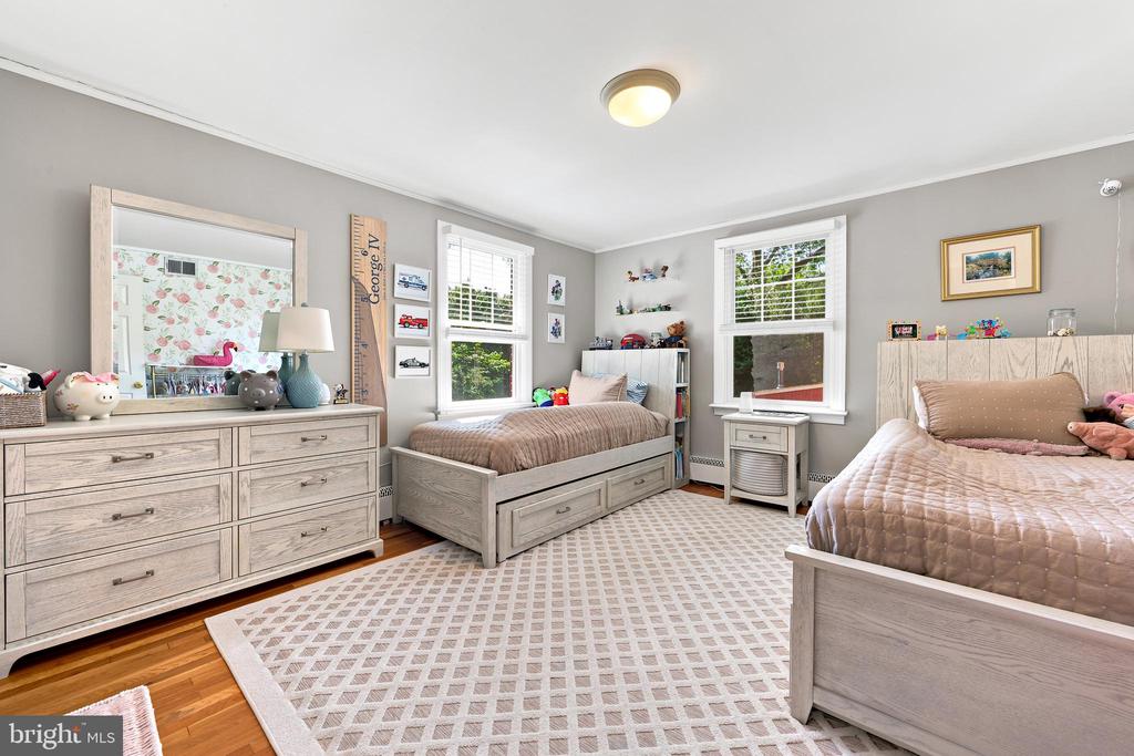 Bedroom with two beds, light wood furniture, a geometric rug, and windows with white blinds.