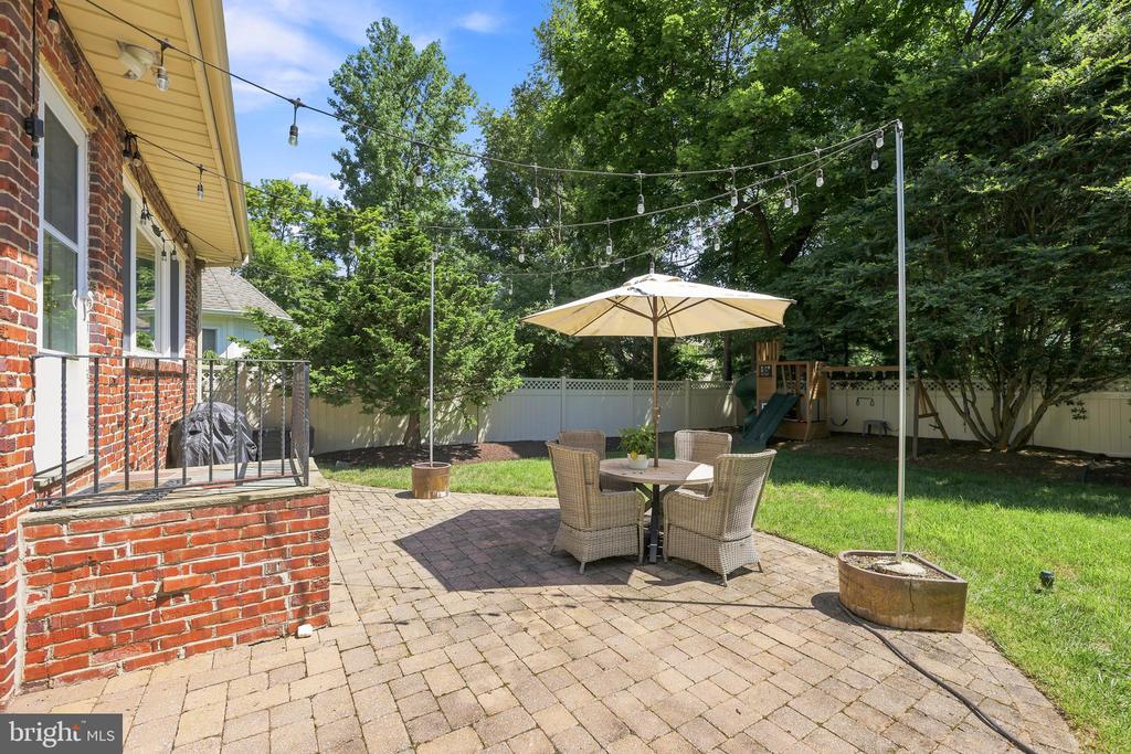 Backyard patio with brick paving, outdoor dining furniture, umbrella, string lights, and mature trees.