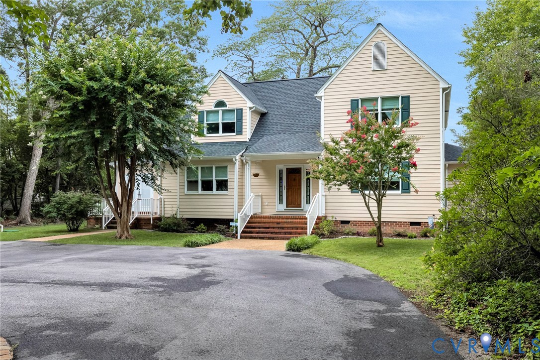 Two-story beige home with green shutters, brick steps, and flowering shrubs beside the driveway.