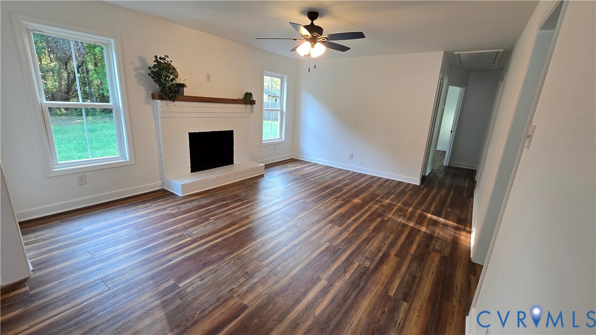 Empty living room with dark wood floors, a white brick fireplace, and a ceiling fan.