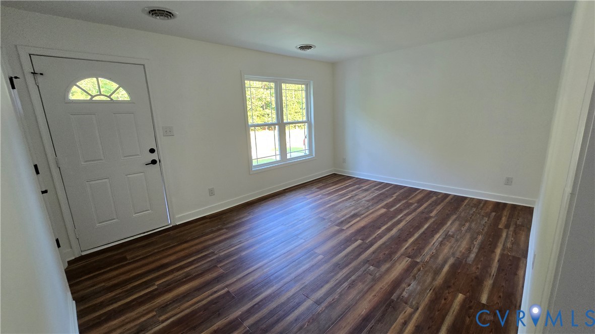 Vacant room with dark plank flooring, a front entry door, and a sunlit window.