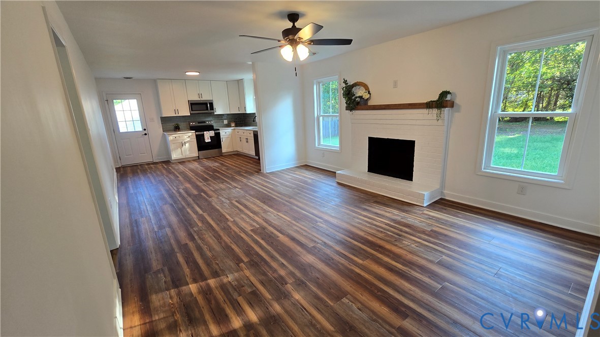 Unfurnished living room with dark wood-look floors, a white brick fireplace, and an open kitchen.