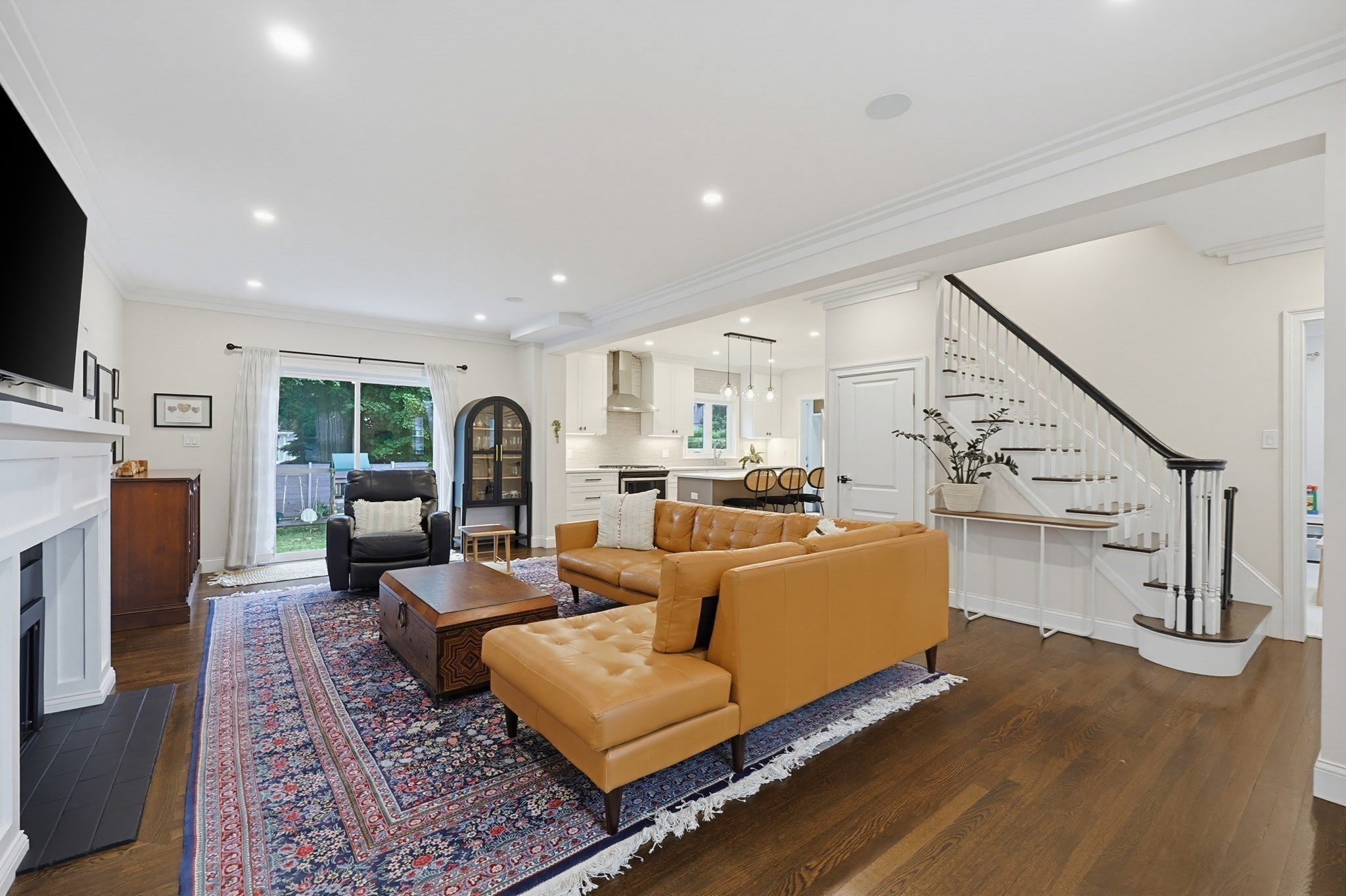 Living room with a caramel leather sectional, patterned rug, white fireplace, and open kitchen.