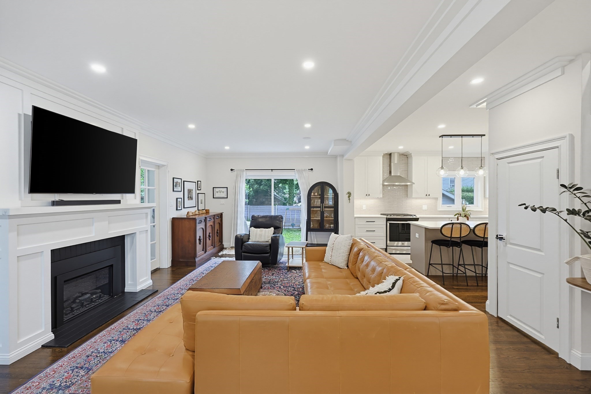 Open-plan living room and kitchen with a tan leather sectional, white fireplace, and island with bar stools.
