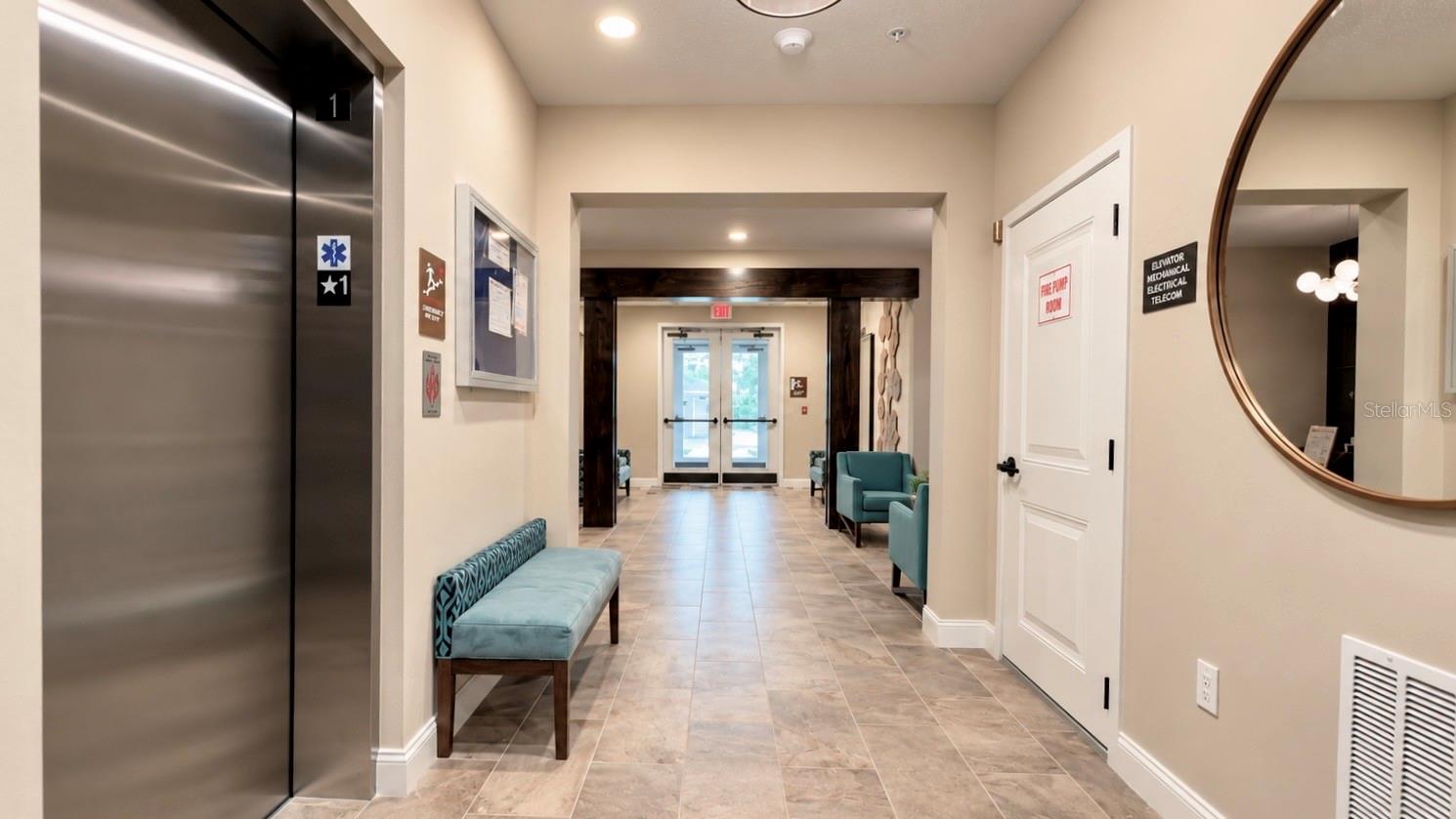 Apartment lobby hallway with a stainless-steel elevator, teal seating, tiled floor, and glass double doors.