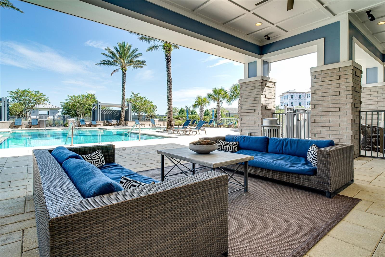 Covered poolside lounge with blue-cushioned wicker sofas and palm trees beside the swimming pool.