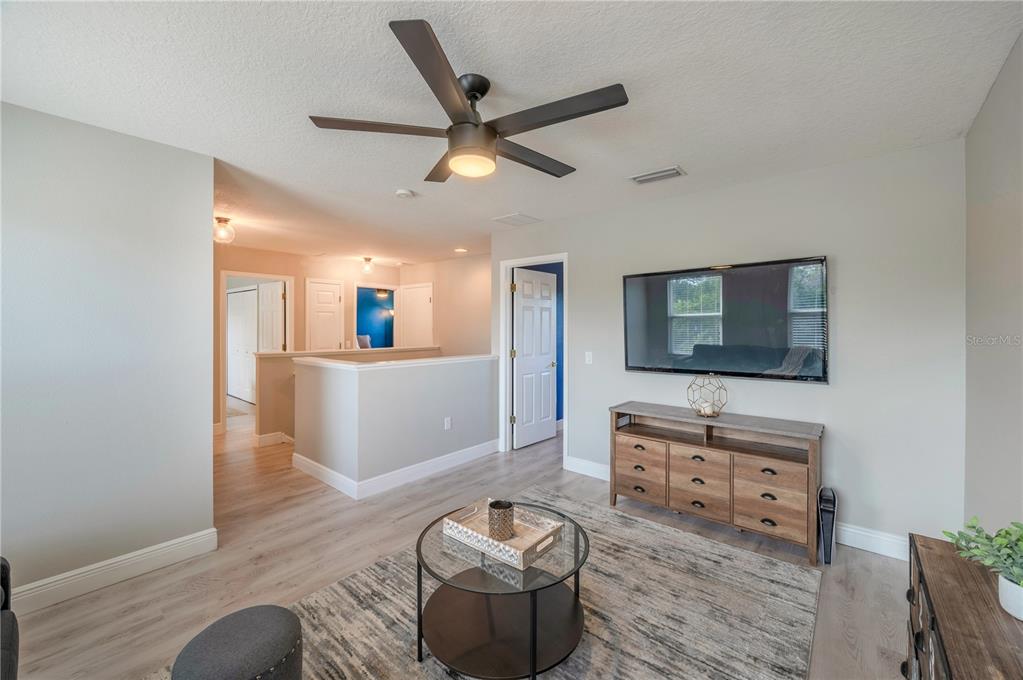 Living room with a ceiling fan, wall-mounted TV, wooden dresser, and glass coffee table on a patterned rug.