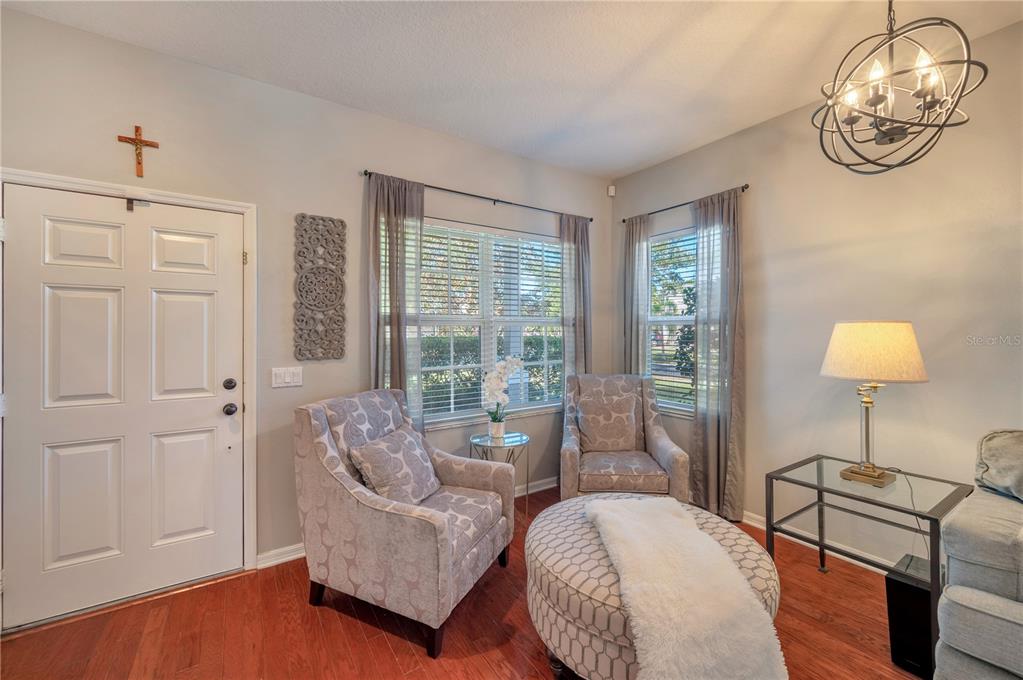 Living room with patterned armchairs, a round ottoman, shuttered windows, and warm wood flooring.