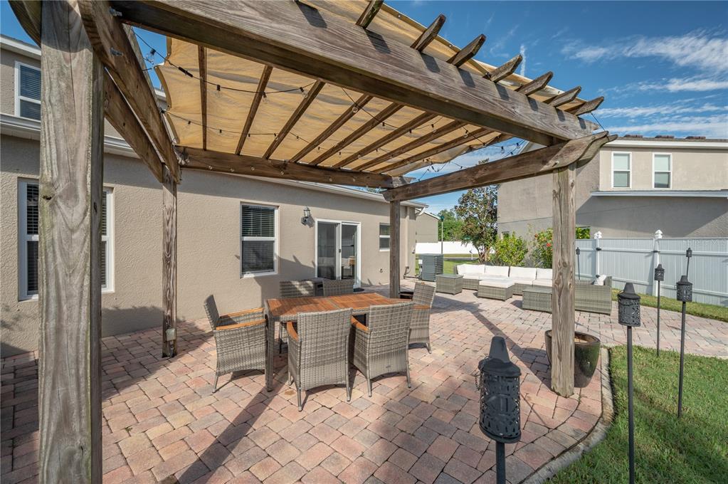 Paver patio beneath a wooden pergola, furnished with a wicker dining set and lounge seating.